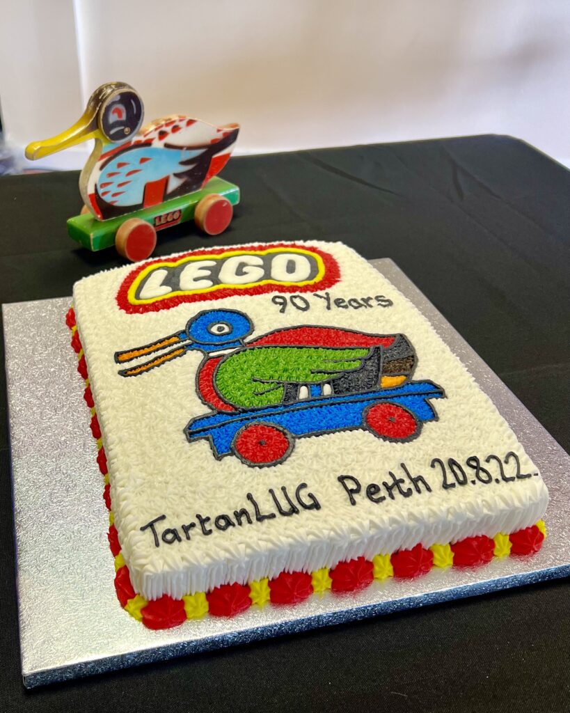 Large cake with classic wooden Lego duck and ’TartanLUG Perth 20.8.22’ decoration. Antique duck toy displayed behind cake.